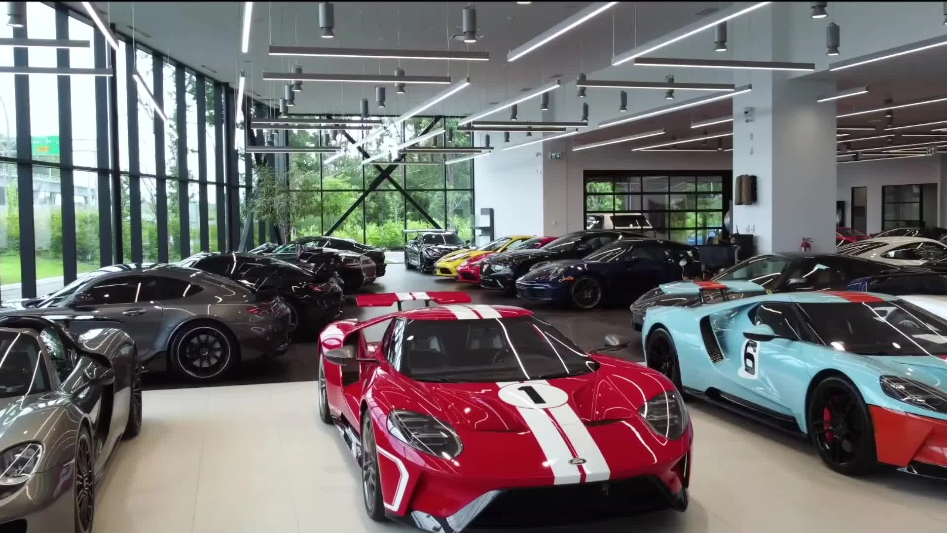 Official John Scotti Luxury-Prestige showroom image with a red sports car bearing white stripes and blue sports cars displayed in a glass-walled gallery.